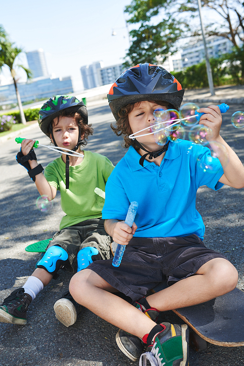 Handsome little brothers in colorful helmets sitting on skateboards and blowing soap bubbles in sunny park