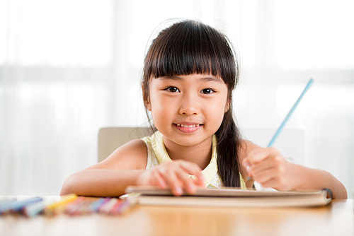 Head and shoulders portrait of adorable Asian girl  with toothy smile while distracted from drawing picture with colorful pencils, white background
