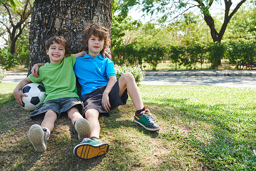 Two joyful little football players  while sitting under tree in sunny green park, full-length portrait
