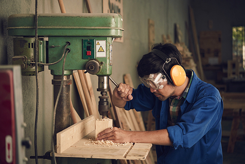 Asian young carpenter using drill press to mae hole in wooden plank