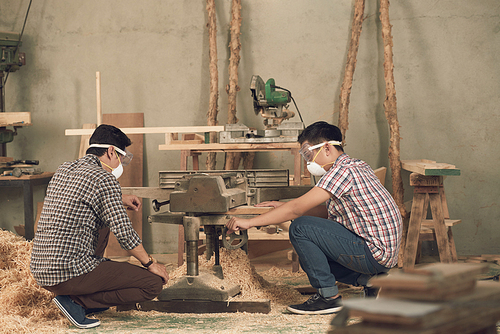 Father and son in masks and goggles using bend saw in workshop
