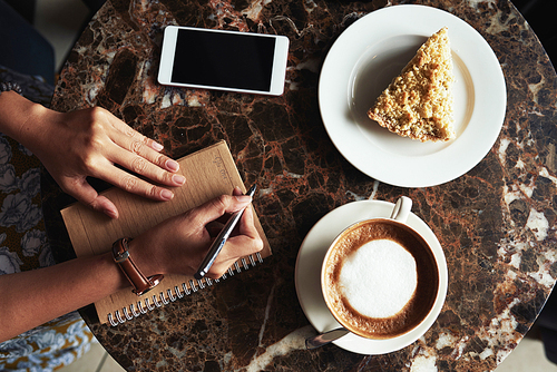 Woman having cappuccino and tart when writing down her plans and ideas