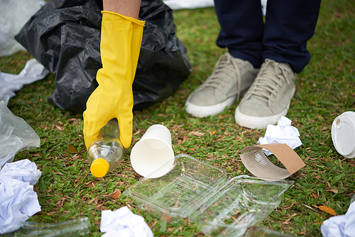 Close-up shot of plastic waste lying on grass, human hand taking bottle in order to put it into bin bag