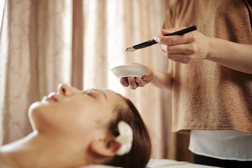 Beautician using small bowl and brush when applying cream mask on face of female patient