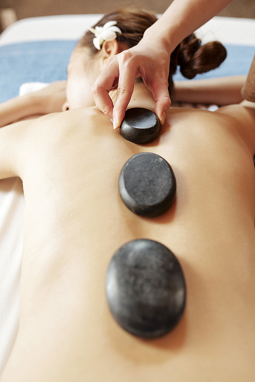 Spa salon worker putting hot basalt stones along spine of young female client