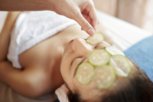 Cosmetologist putting cucumber slices on face of young pretty Asian woman