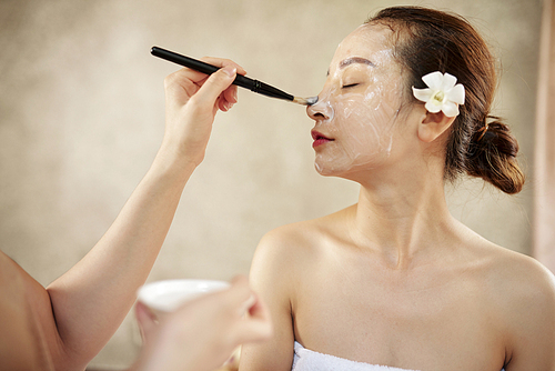 Beautician applying creamy face mask on face of beautiful Vietnamese woman