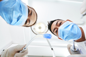 Low angle portrait of male doctor and his assistant wearing protective masks using special instruments to treat patient's teeth in clinic