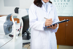Female ophthalmologist standing in white coat and writing a prescription on clipboard in ophthalmology clinic