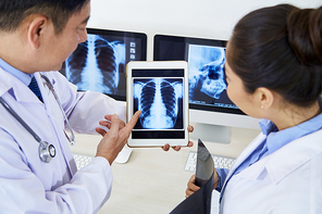 Mature male radiologist holding tablet pc with x-ray image of lungs and discussing the problem with his assistant at hospital