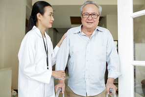 Old man in eyeglasses standing with walker and  while nurse in white coat helping him to move