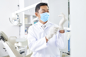 Mature male dentist in uniform and protective mask on his face wearing gloves and is going to treat his patient in clinic