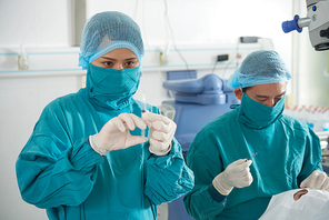 Two surgeons in uniform working in operating room with patient, female nurse standing with syringe while surgeon making an operation on patient's eyes
