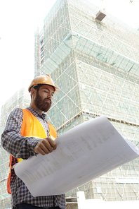 Serious foreman examining blueprint of skyscraper his company building