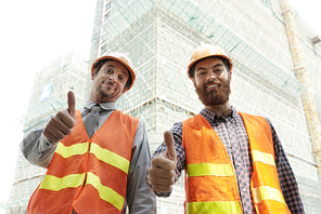 Cheerful construction site workers in bright vests showing thumbs-up and smiling at camera