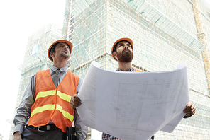 Two professional engineer rolling out paper blueprint when looking at skyscraper