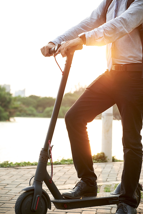 Close-up of young businessman standing on electric scooter and riding on it in the park after work outdoors