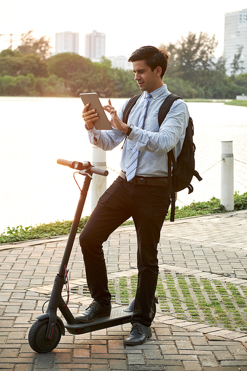 Young office worker with backpack behind his back using digital tablet and communicating online during his riding on scooter in the park