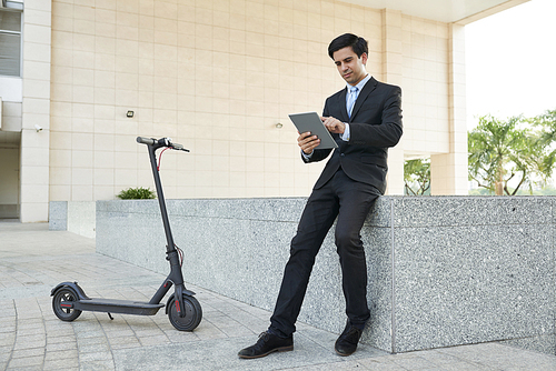 Young businessman in suit working online on digital tablet while sitting in the city with electric scooter