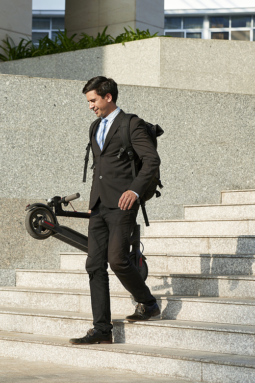 Young businessman in suit with backpack behind his back holding electric scooter in his hand and walking down the stairs in the city