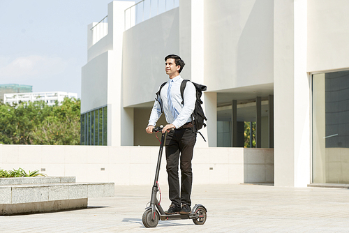 Young smiling manager with backpack behind his back riding on electric scooter past the office building outdoors