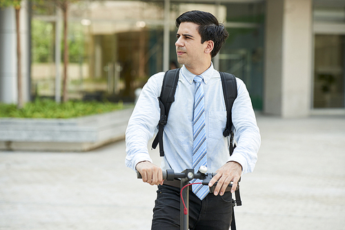 Young office worker with backpack behind his back standing on scooter and looking away in the city outdoors