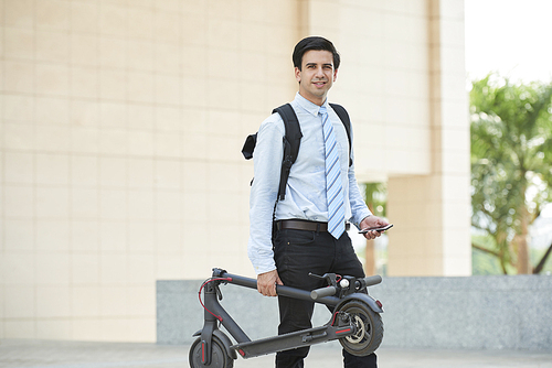 Portrait of young handsome businessman with backpack behind his back holding scooter in one hand and mobile phone in another and smiling at camera outdoors