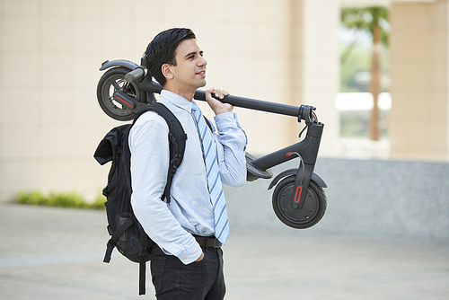 Young handsome businessman with backpack on his back standing with scooter and looking away while riding in the city