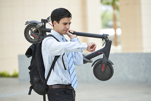Young businessman standing with backpack behind his back holding electric scooter and looking at the time on watch on his hand while standing in the city.