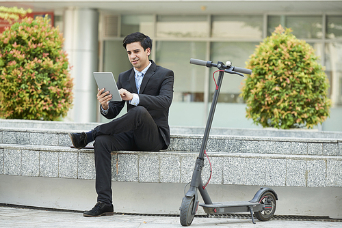 Young businessman in suit sitting in the city and using digital tablet after riding on scooter outdoors