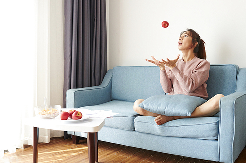 Asian young girl throwing a fresh red apple while resting on sofa during meal time in the living room at home
