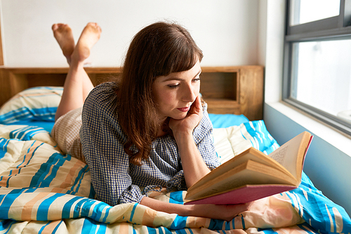 Pretty young woman lying on bed in her room and reading a book