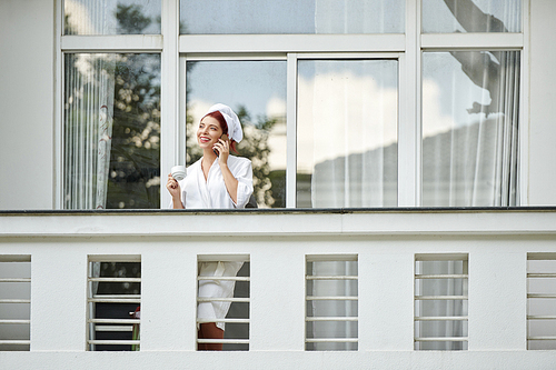 Pretty happy woman in bathrome standing on balcony of her hotel room and talking on phone