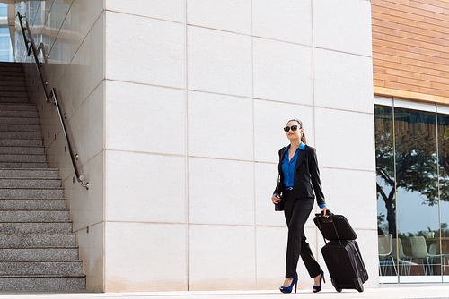 Businesswoman in glasses and suit walking in the street with suitcase