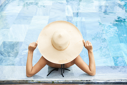 Young woman in big straw hat relaxing in swimming pool with turquoise water, view from above