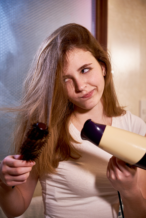 Young woman brushing her long hair with hairbrush and drying with hairdryer in bathroom