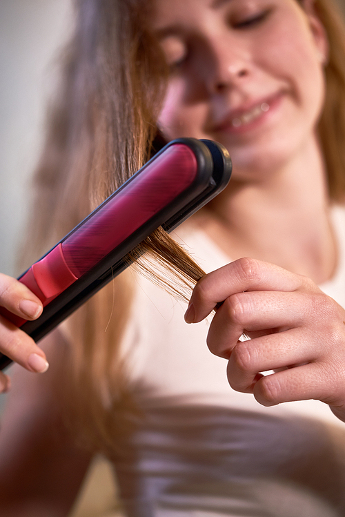 Close-up of young woman holding hair straightener and straightening her long hair herself