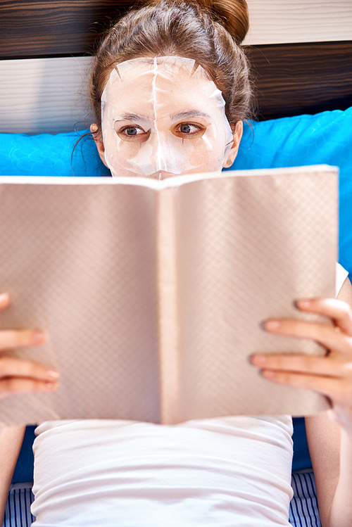 Young woman applying paper facial mask on her face and reading a book in bed