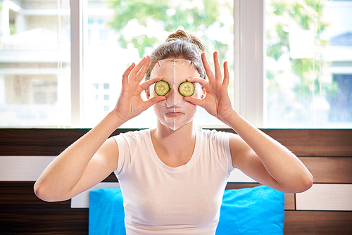 Portrait of young woman applying facial mask on her face and holding slices of cucumbers in front of her eyes sitting in bedroom