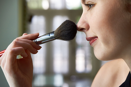 Close-up of young woman applying dry cosmetic foundation on her face using makeup brush