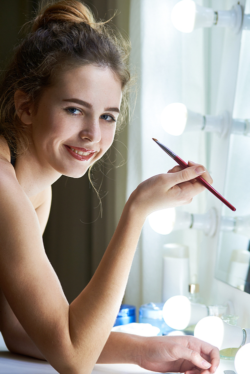 Portrait of young beautiful woman standing in front of the mirror with cosmetic brush and smiling at camera
