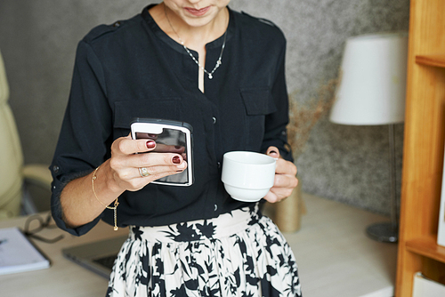 Businesswoman sitting on edge of her tablet drinking cup of coffee and reading messages in her phone