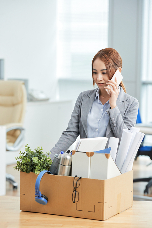 Asian female employee standing near the cardboard box with office stuffs and talking on mobile phone at office