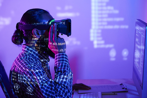 Modern programmer in vr headset sitting in front of computer monitor while coded data reflecting on him