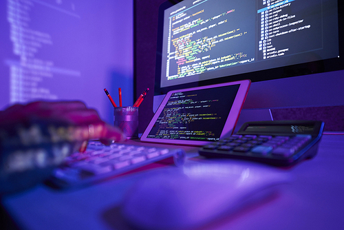 Close-up of busy man sitting at the table in front of computer monitor and digital tablet typing and coding in dark office