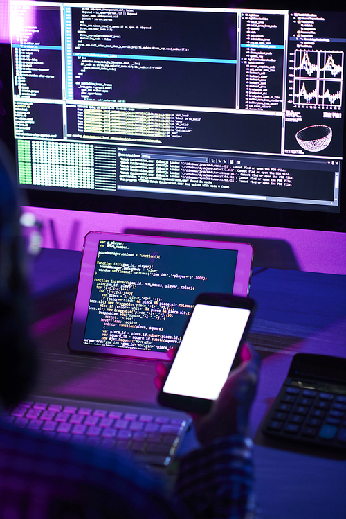 Modern programmer with smartphone sitting by workplace in front of tablet and computer monitor with coded data