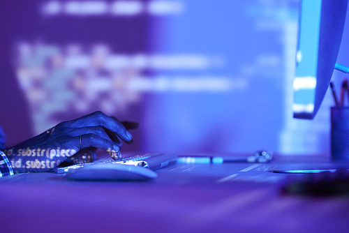 Close-up of young developer sitting at office desk and typing on computer keyboard in dark office