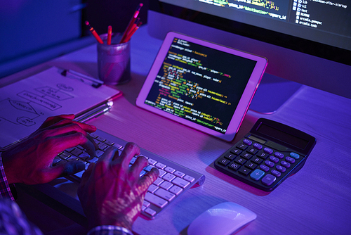 Close-up of man sitting at the table in front of tablet pc with software and typing on computer keyboard in dark office