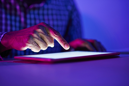 Close-up of young man sitting at the table and touching the display of digital tablet in dark room