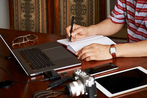 Close-up of photographer sitting at the table among wireless gadgets and writing a list of new orders from his clients in notepad
